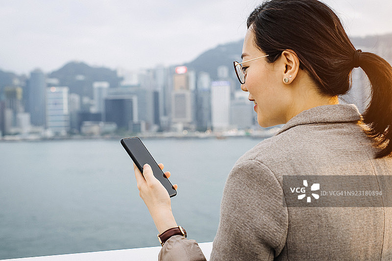 香港维多利亚港，一名微笑的年轻女子用智能手机给客户发送电子邮件图片素材
