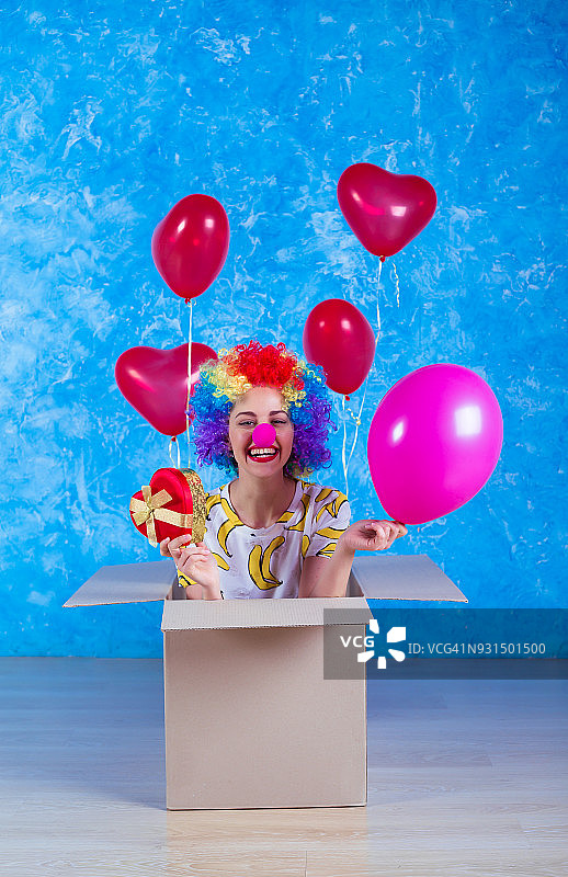 1愚人节的概念。滑稽的小丑坐在一个带着氦气球的纸箱里，看着镜头。生日礼物。图片素材