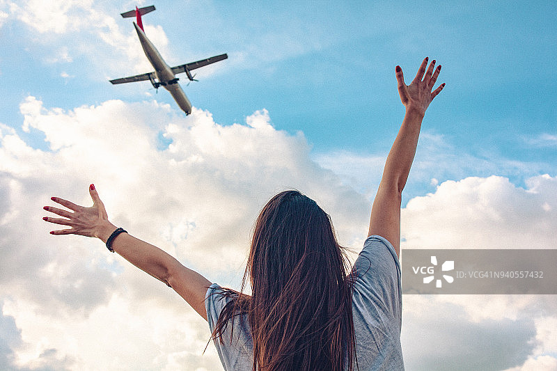 一个年轻女子的后视图与伸出的双臂挥手向飞行的商业飞机上方她图片素材