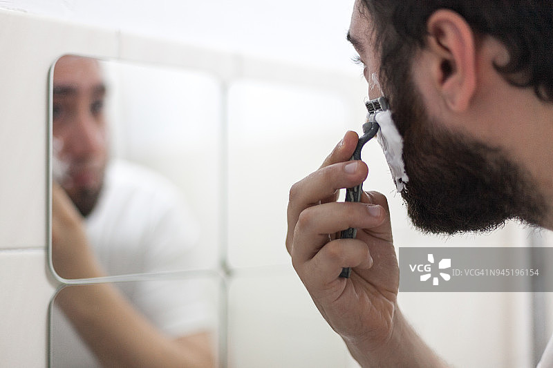 一名男子拿着剃须刀在镜子前刮脸，脸上有泡沫图片素材