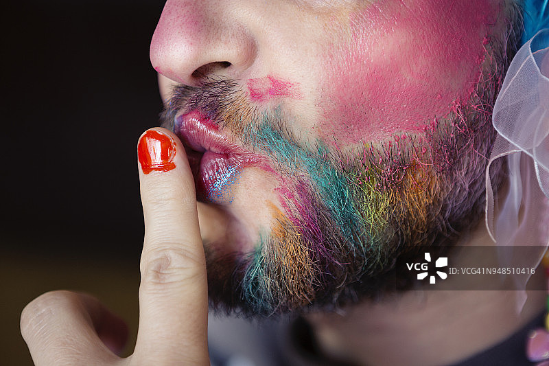 一个留着胡子的男人，画着五颜六色的妆，涂着指甲油，同时把手指放在嘴唇上图片素材