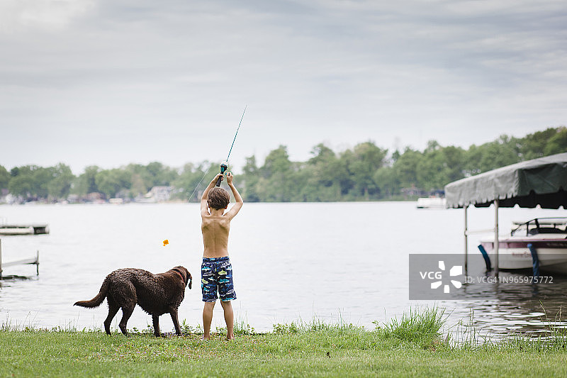 后视图赤膊男孩钓鱼，而站在湖边的狗对多云的天空图片素材
