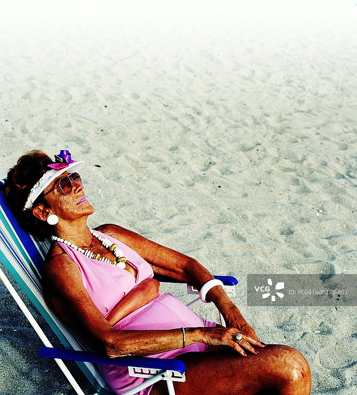 穿着粉红色比基尼的老妇人，坐在沙滩上的帆布躺椅上图片素材