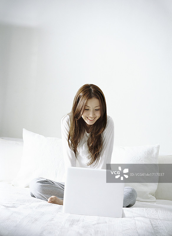 年轻女子盘腿坐在床上使用笔记本电脑图片素材
