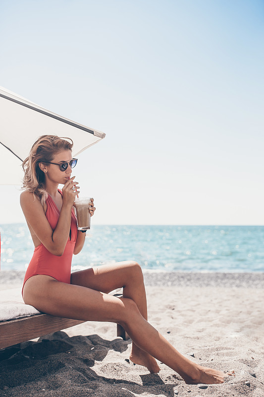 年轻女子拿着鸡尾酒杯坐在白色沙滩上的日光浴床上图片下载