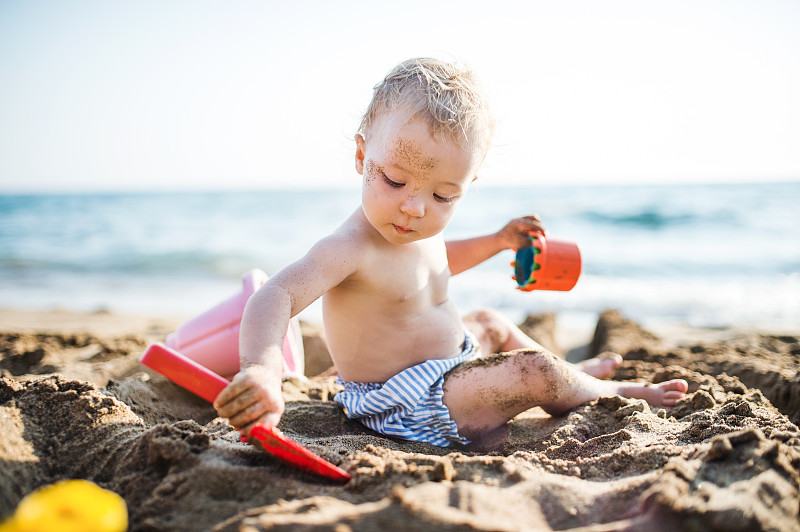 暑假，一个蹒跚学步的小女孩坐在海滩上玩耍。图片素材