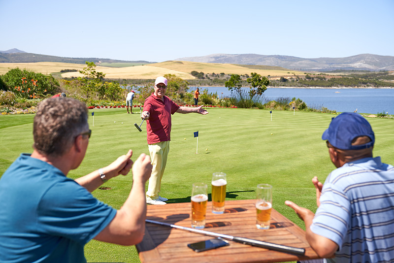 快乐的男性高尔夫球手喝着啤酒，在高尔夫球场练习推杆图片下载
