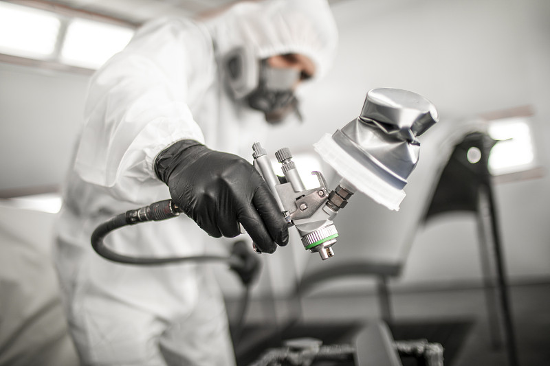 车身车间的技术员在一个喷漆室里，穿着全套工作服和一个呼吸面罩，在一个汽车部件上涂抹一层油漆图片下载