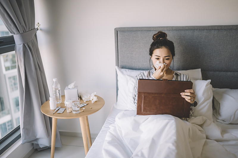 一位亚洲华裔女性在床上穿着睡衣，用纸巾捂着鼻子看数字平板电脑图片下载