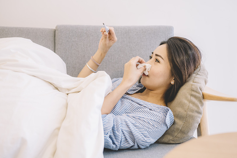 一名亚洲华裔女性躺在沙发上，穿着睡衣和毯子看着温度计图片下载