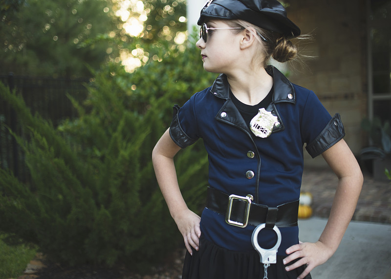 万圣节前夕，一个自信的女孩穿着警服，双手放在臀部，靠着院子里的植物站立图片下载