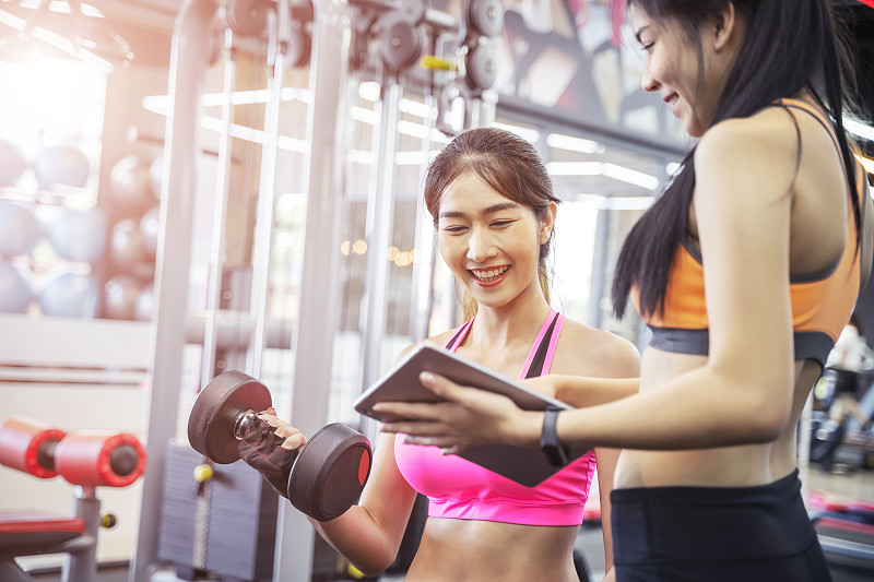 年轻的亚洲女子在健身房和她的私人教练锻炼。私人教练帮助妇女使用沉重的哑铃。图片下载
