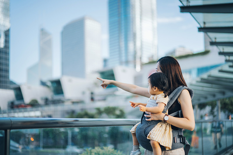 年轻的亚洲母亲指着她的小女儿，在市中心的城市露台上对着现代摩天大楼图片下载