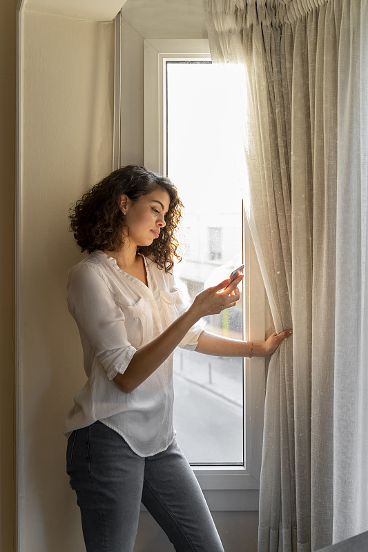 一个年轻女子在家里窗户边用手机图片下载