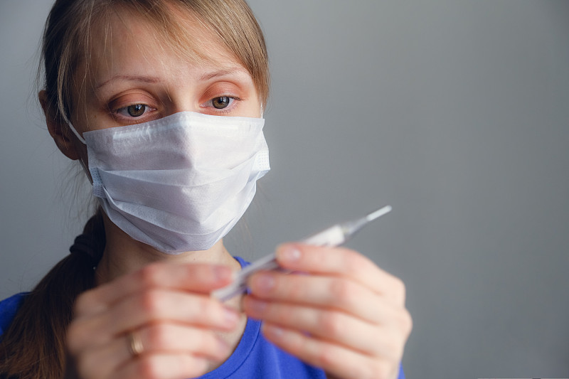 健康的概念。一个生病的女孩戴着一次性医用口罩看温度计。关闭。女孩检查了体温。流感季节，流感病毒。削弱了免疫力。图片素材