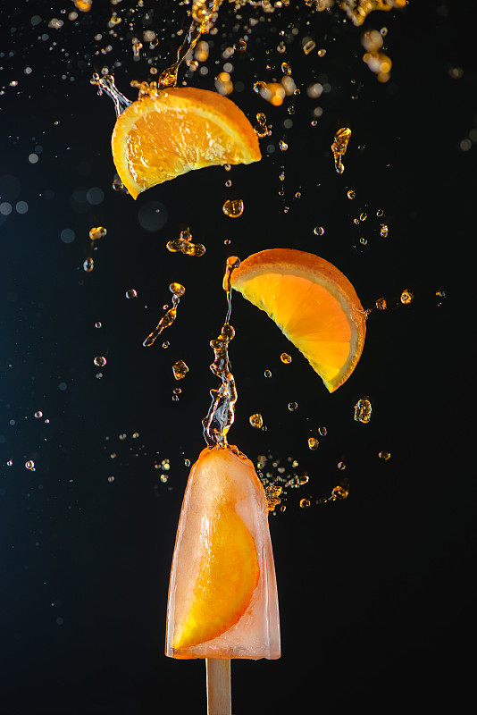 冰棒与新鲜的橙子片在一个黑暗的背景，动态拍摄与行动，飞溅，水滴和散焦。图片素材