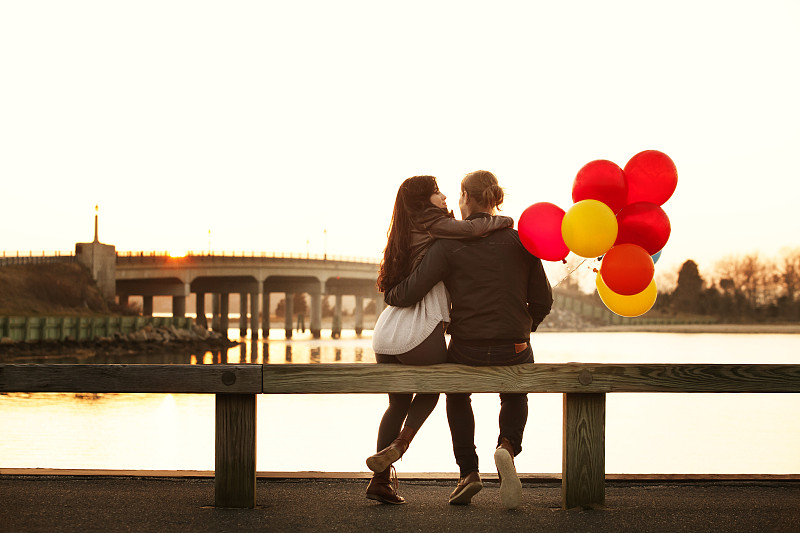 在日落期间，气球坐在栏杆上的夫妇的后视图图片下载