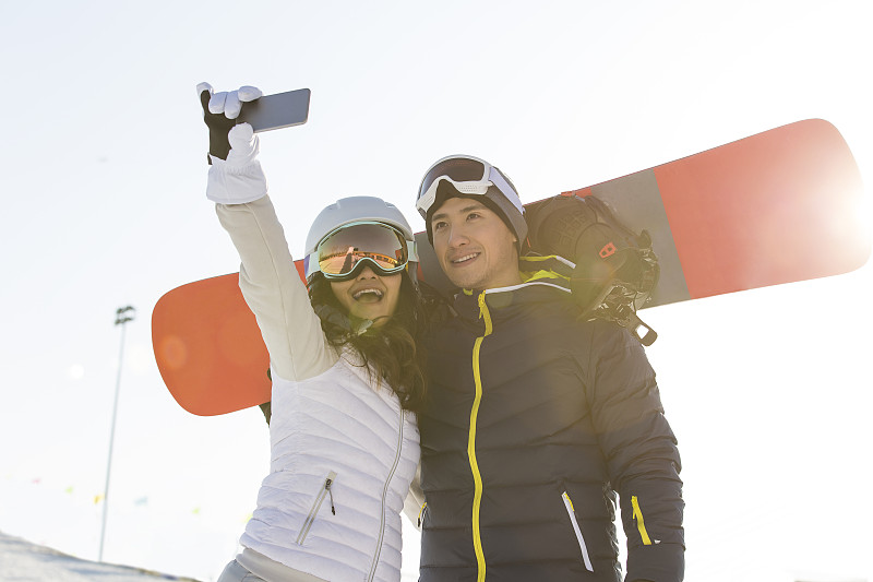 甜蜜情侣背着滑雪板在户外滑雪场拿手机自拍图片下载