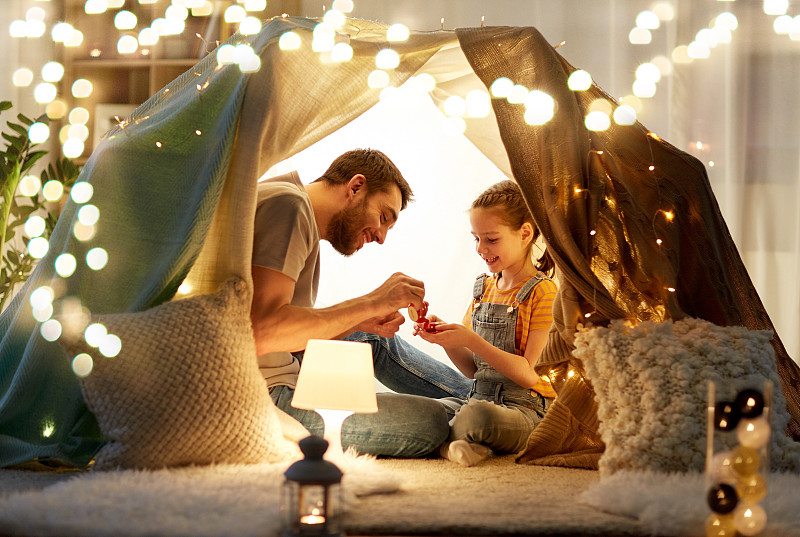 家庭、hygge和人们的概念——快乐的父亲和小女儿晚上在家里的孩子帐篷里玩茶会。家庭在孩子们的帐篷里玩茶话会图片素材