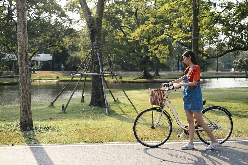 长发女孩穿着橙色t恤和牛仔裤套头衫，骑着自行车沿着公园的草地走。图片下载