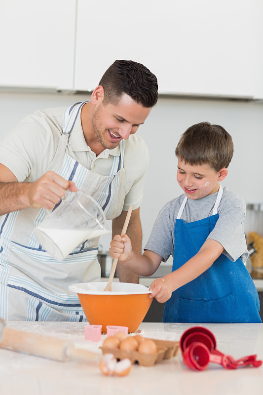 快乐的父亲倒牛奶在碗里，与儿子在厨房柜台上烤饼干图片下载