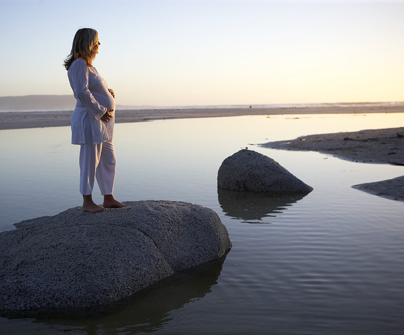 孕妇站在沙滩上的岩石上，双手抱着肚子图片下载