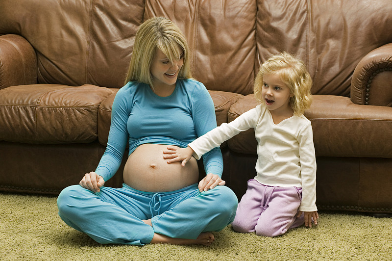 女儿(4-5岁)坐在沙发旁的地板上抚摸着孕妇的肚子图片下载