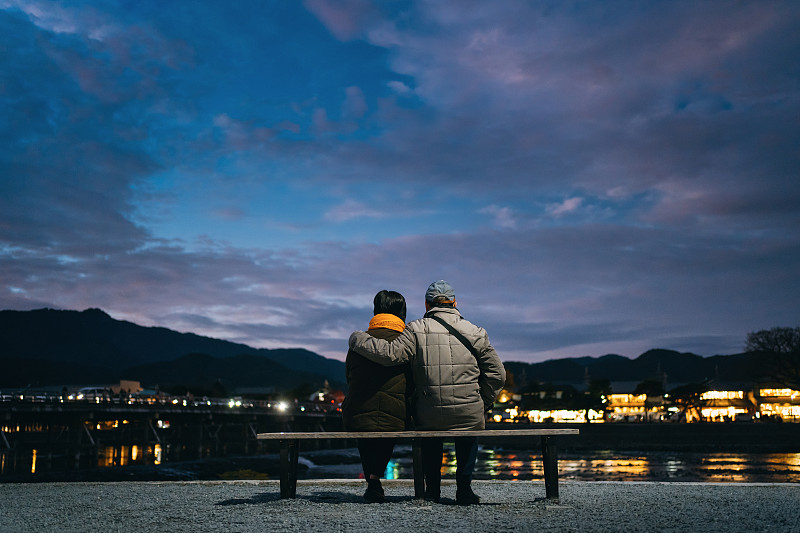 后视图的资深夫妇坐在长椅上并排欣赏日落的湖，而访问传统日本城镇东山，京都，日本黄昏图片下载