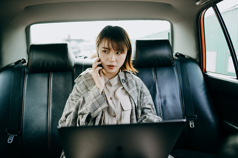 年轻的亚洲女商人坐在出租车后座，一边用智能手机聊天，一边用笔记本电脑工作图片素材