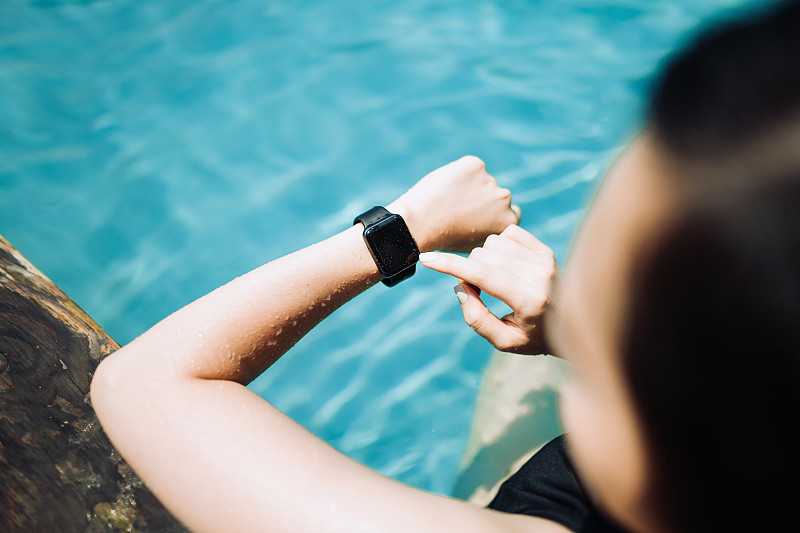 头顶上的年轻女子使用智能手表，同时享受泳池边的阳光图片素材