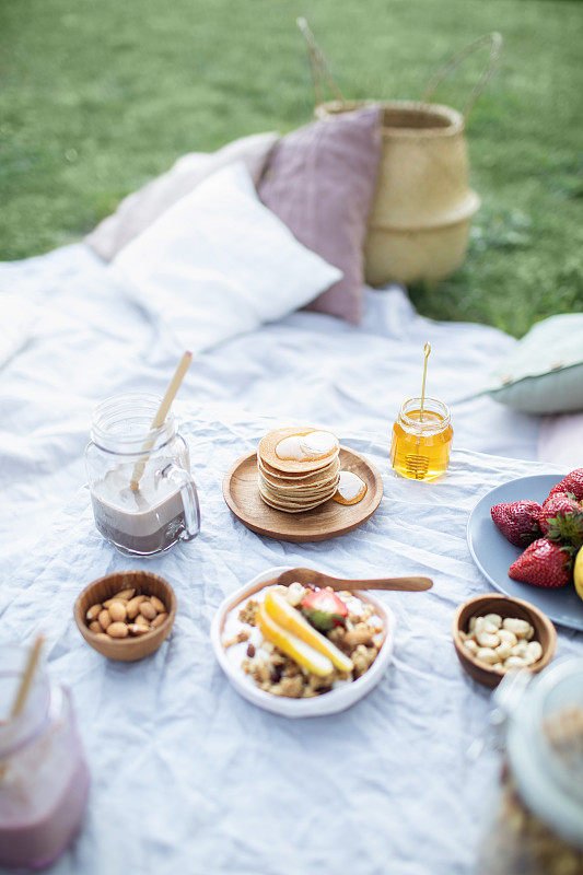 夏季健康素食野餐在公园。
水果，可可，蜂蜜煎饼和有机格兰诺拉麦片在亚麻桌布上装饰枕头和柳条篮子。图片下载