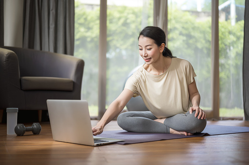 一个女人在家里用笔记本电脑做瑜伽，在网上找到锻炼教程。家庭隔离期间的家庭健身直播。健康的生活方式和工作生活的平衡理念。图片素材
