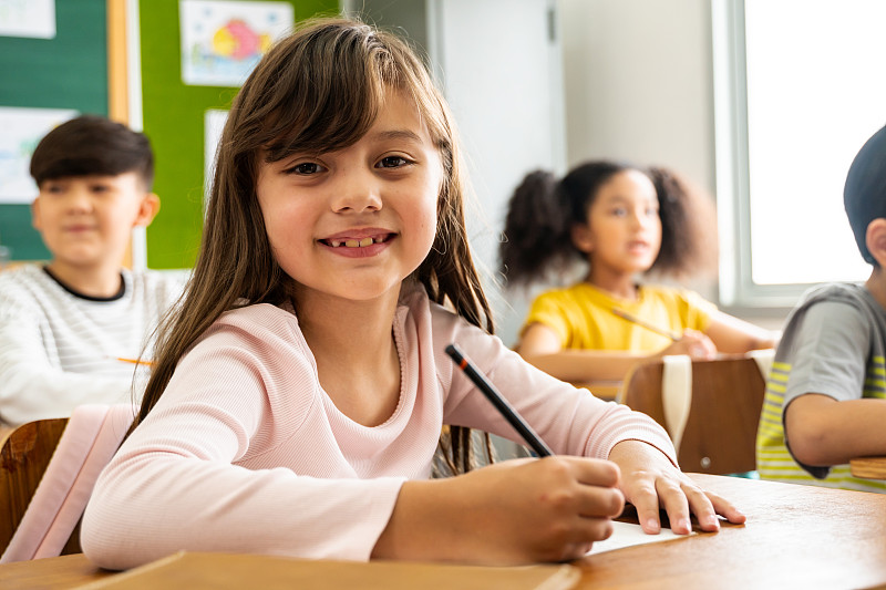 快乐的女学生坐在课桌上微笑的肖像。可爱的白人小学女生坐在学校的纸上写字，看着相机，教育，学习，幸福图片素材