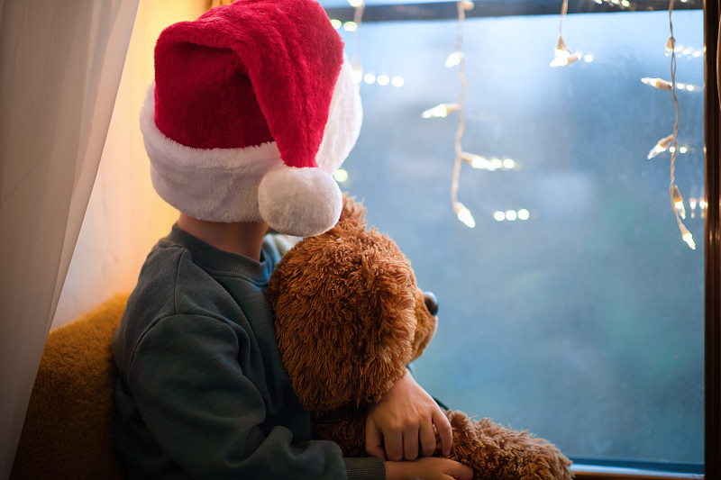 可爱的小男孩在家庭隔离戴着圣诞帽和他的泰迪熊在圣诞节图片下载