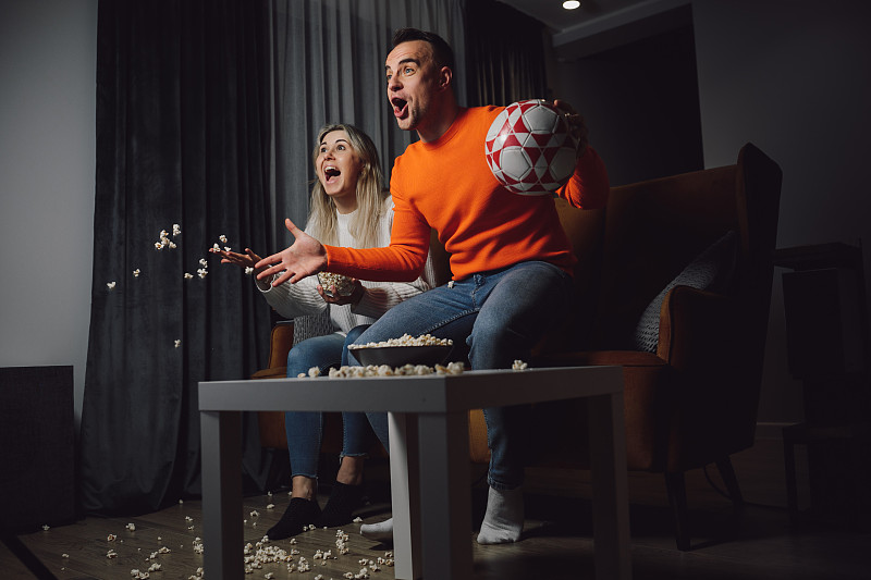 黑头发站着跳跃的热情的男人和金发尖叫的女人在沙发上看电视上的足球胜利进球图片下载