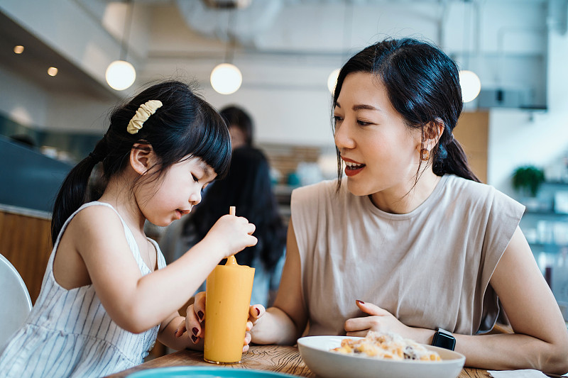年轻的亚洲母亲和可爱的小女儿一起在咖啡馆吃午餐，分享芒果冰沙和意大利面。他们在交谈，看着对方，开心地笑着。母女关系。家庭和外出就餐的生活方式图片下载