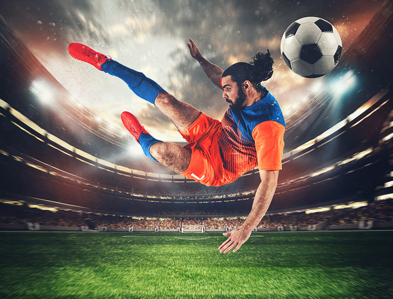身穿橙色和蓝色制服的足球前锋在体育场用一记杂技般的踢腿将球踢向空中图片下载