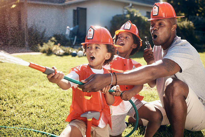 穿着消防员服装的小男孩。一位非裔美国父亲正在和他的孩子们玩耍。小男孩在花园里玩。男孩们在外面用水管喷水。兴奋的兄弟玩图片下载