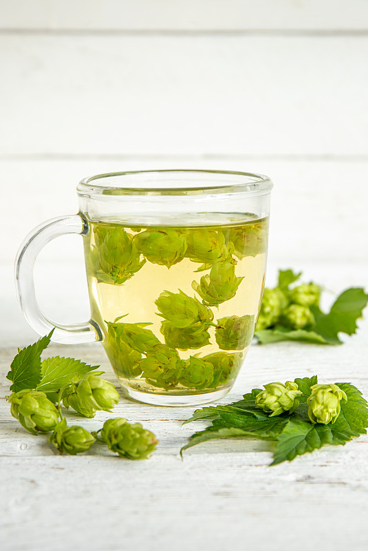 凉茶饮料是由葎草制成的，俗称啤酒花。啤酒花与茶杯在白色的木材背景，在家里的内部。图片下载