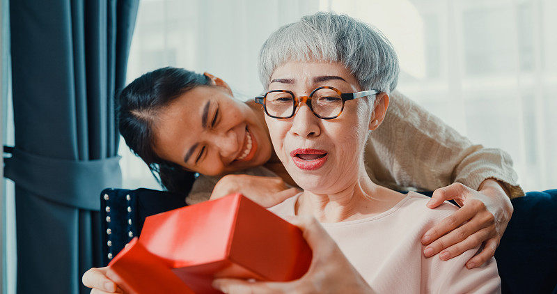 快乐的亚洲女儿惊喜礼物生日礼物在红色礼盒退休资深母亲坐在沙发沙发在家里。图片下载