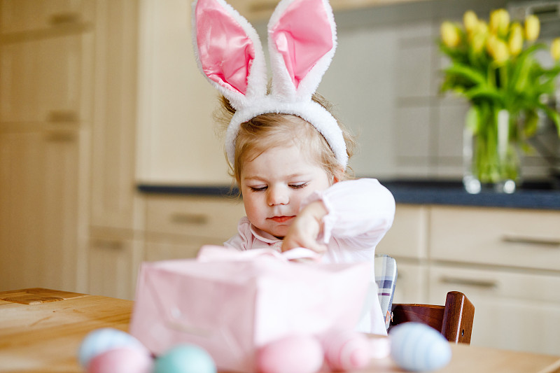 可爱的小蹒跚学步的女孩戴着复活节兔子耳朵玩彩色粉彩蛋。快乐的宝宝孩子拆礼物。可爱的健康微笑的孩子在粉红色的衣服享受家庭假期图片下载