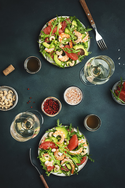 海鲜沙拉配虾仁、鳄梨、葡萄柚、芝麻菜和腰果。餐桌上摆放着白葡萄酒。深绿色背景，俯视图图片下载