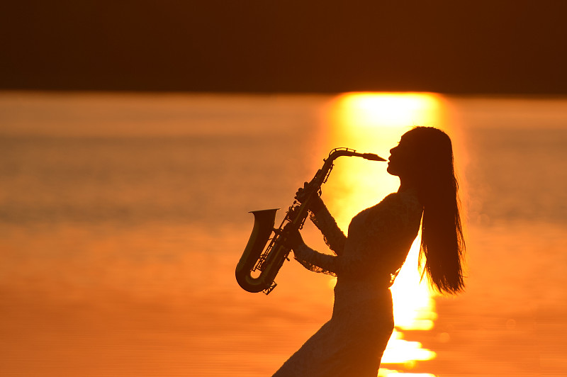 夕阳下的泰国，一名女萨克斯手在湖边吹奏萨克斯图片下载