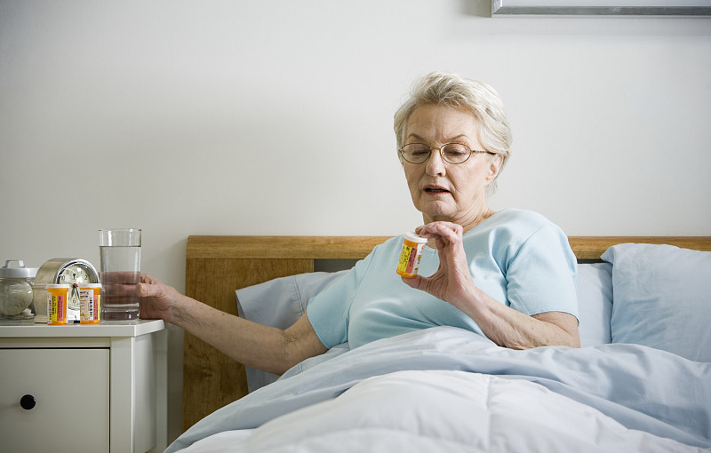 老妇人斜倚在床上，手里拿着一杯水和一瓶药图片下载