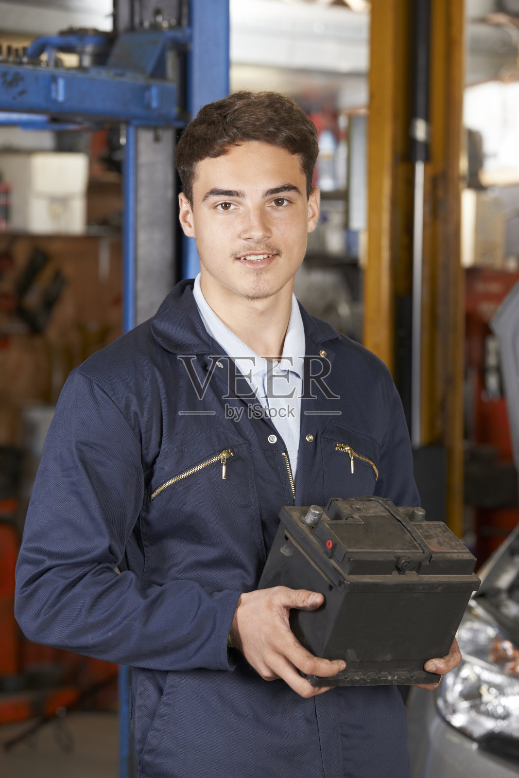 在汽车修理厂拿着汽车电池的学徒机械师照片摄影图片