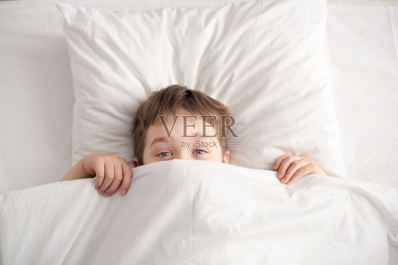 快乐的男孩躺在白色的床上，盖着白色的毯子照片摄影图片