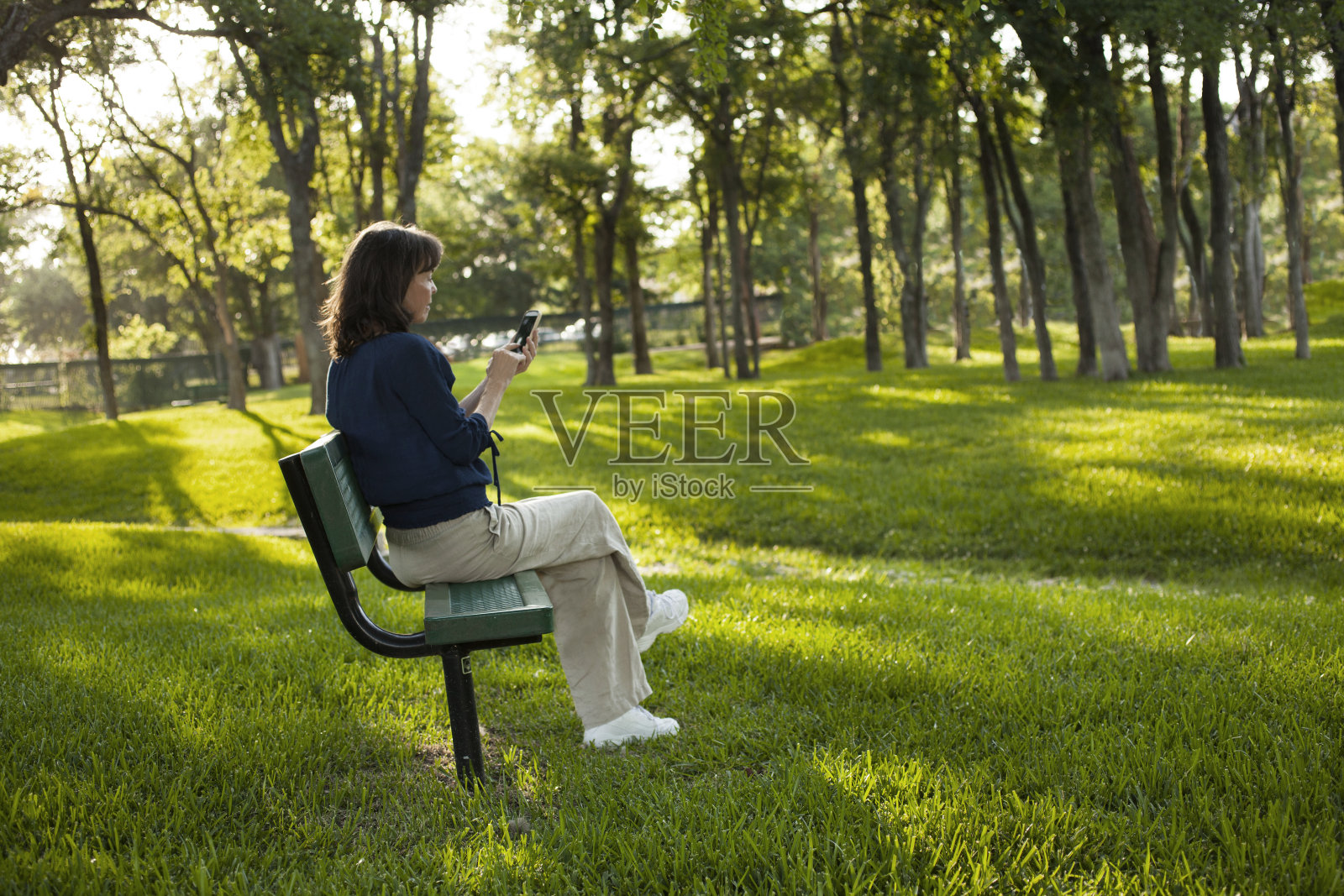 坐在公园长椅上的成熟女人。在夏天发短信。照片摄影图片