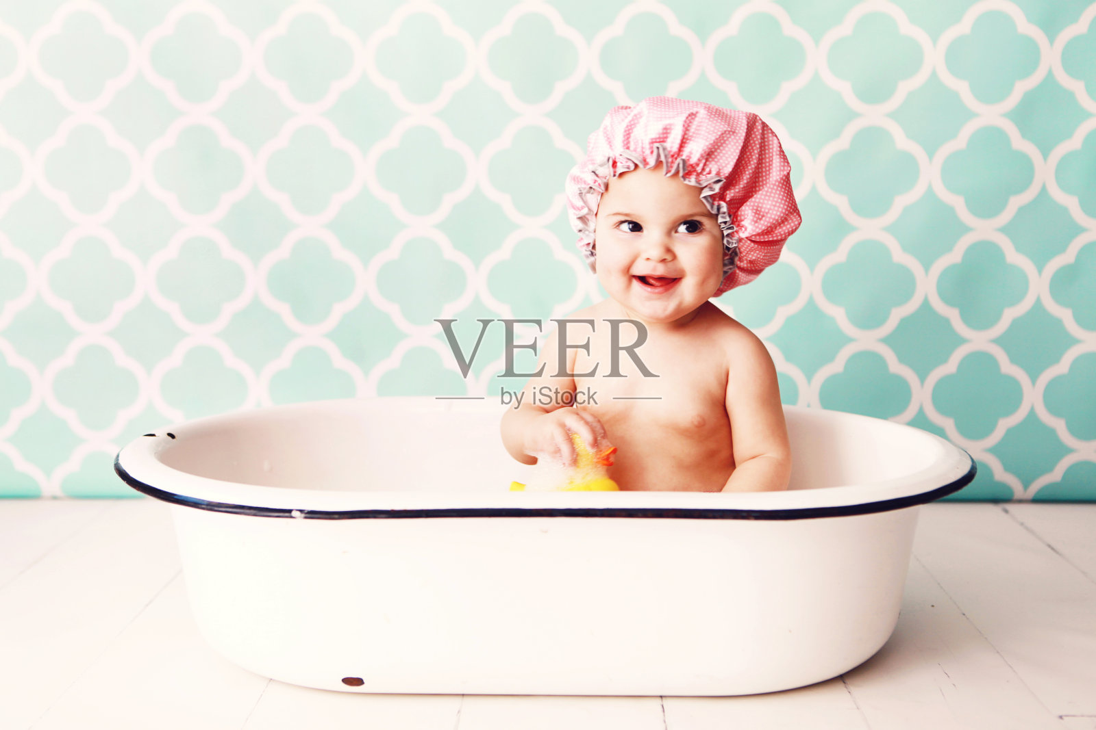 戴着粉色浴帽的宝宝在浴缸里玩橡皮鸭照片摄影图片