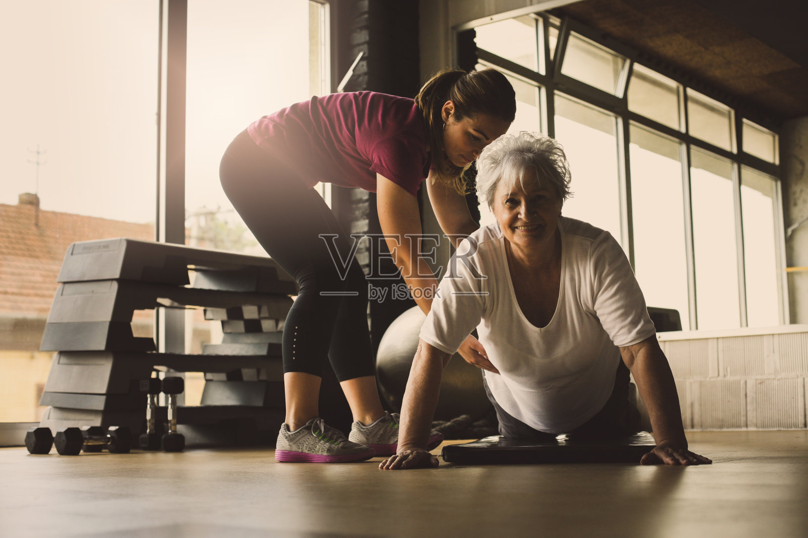 做俯卧撑的老女人。年轻的私人教练帮助高级女性。在康复中心锻炼。看相机。照片摄影图片
