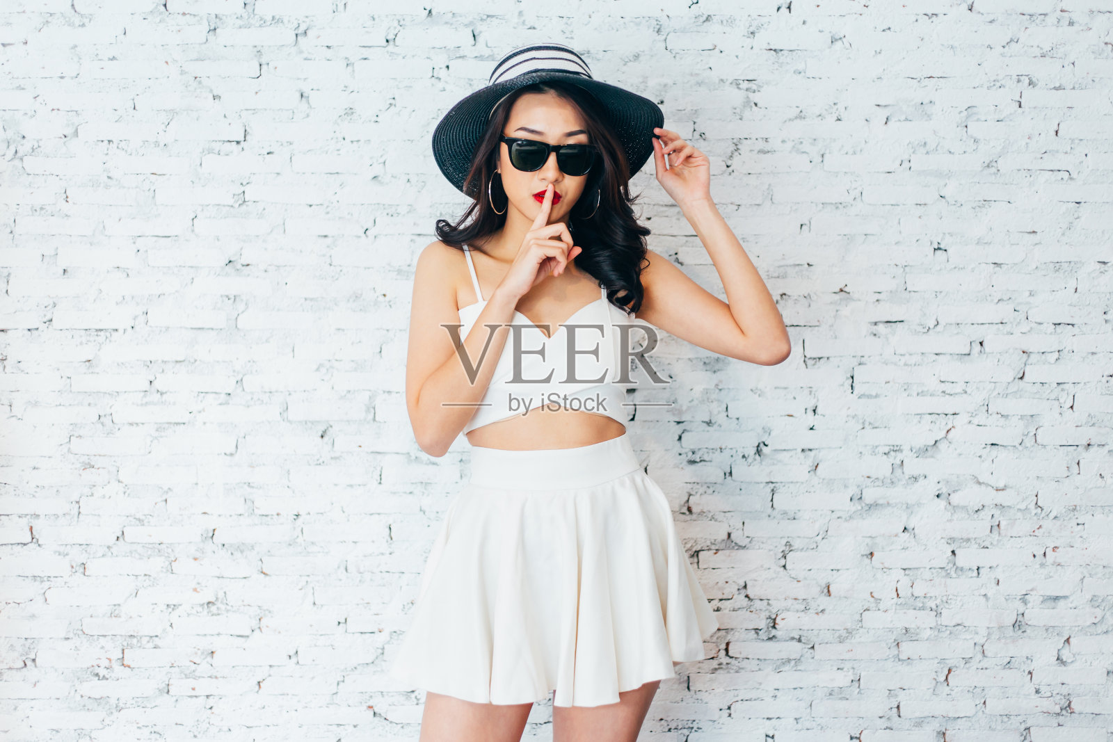 亚洲妇女的手指在她的嘴唇在时尚的服装和夏天的帽子在白色砖墙-沉默嘘手势照片摄影图片